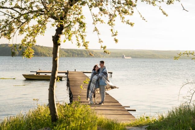 A couple standing on a wooden dock by a lake, wrapped in a blanket, with a small boat tied to the dock and a serene lake in the background, capturing a peaceful and romantic moment during their anniversary trip.