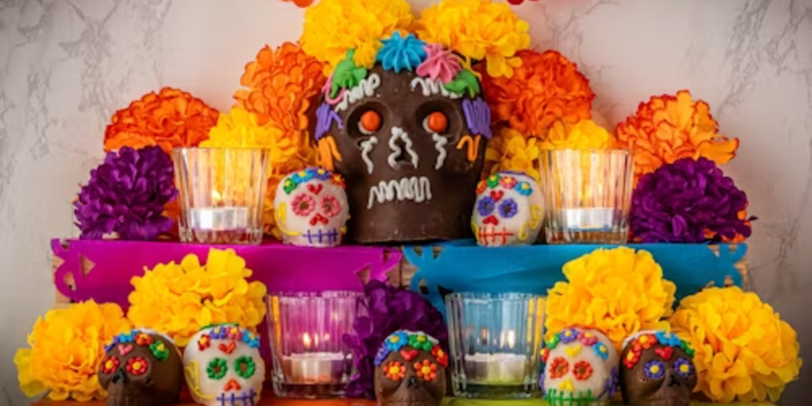 A vibrant Día de los Muertos altar adorned with colorful marigold flowers, decorated sugar skulls, and lit candles. A large chocolate skull with intricate icing decorations sits at the center, surrounded by smaller skulls with bright floral patterns. The altar is set against a white wall, creating a festive and traditional tribute.