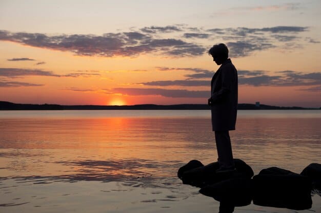 A silhouette of a person standing on rocks by a calm lake during a vibrant sunset. The sky is painted with hues of orange, pink, and purple, reflecting beautifully on the water's surface. The scene is serene and peaceful, capturing the tranquility of nature at dusk.