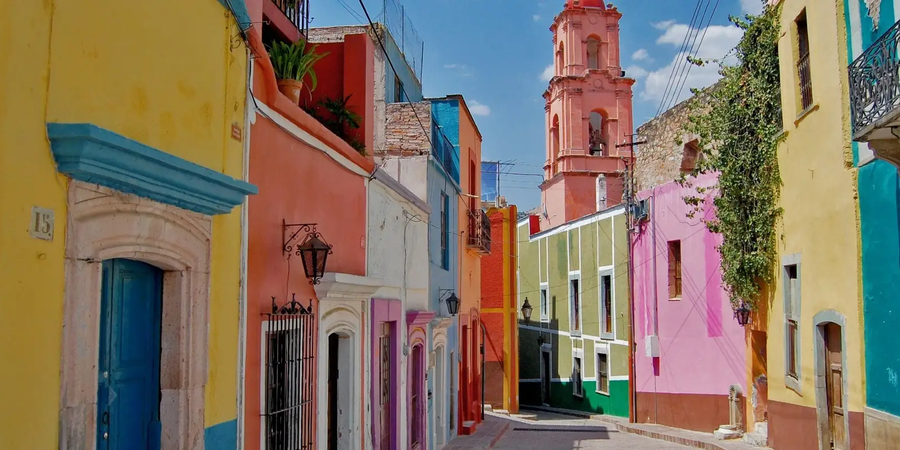 A picturesque street in a historic town with colorful colonial-style buildings, a bright blue sky, and a towering pink bell tower in the background. The narrow cobblestone street is lined with vibrant facades in shades of yellow, pink, green, and blue, adorned with ornate doorways, balconies, and lush greenery.