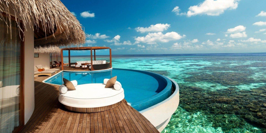 Luxury Maldives overwater villa with infinity pool overlooking crystal-clear turquoise ocean, wooden deck with lounge seating, and thatched-roof design under a bright blue sky.