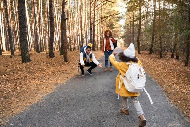 A family is walking in a forest during autumn. The image shows a child in a yellow coat and white hat running towards two adults, one crouching with open arms and the other standing behind. The path is surrounded by tall trees with fallen leaves covering the ground. The scene captures a joyful and heartwarming moment of family bonding in nature.