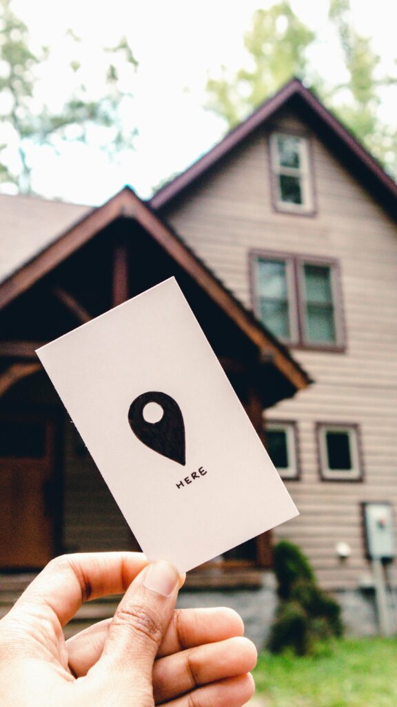 Close-up of card with location icon and the word 'HERE' held in front of a house, suggesting navigation or real estate