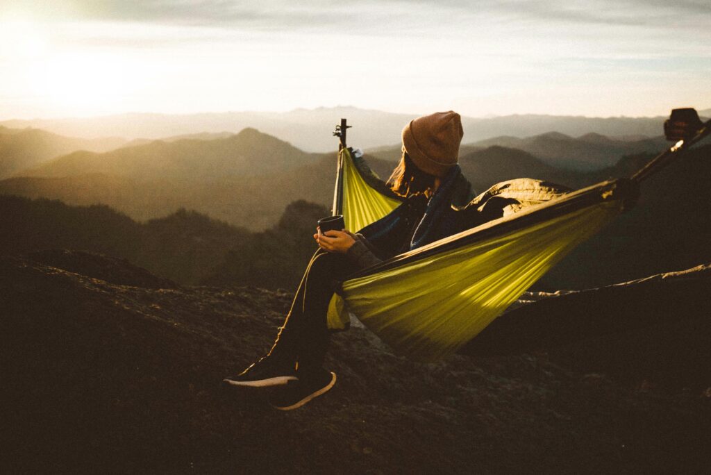 Woman relaxing in a hammock with a hot drink amidst majestic mountain scenery at sunset.