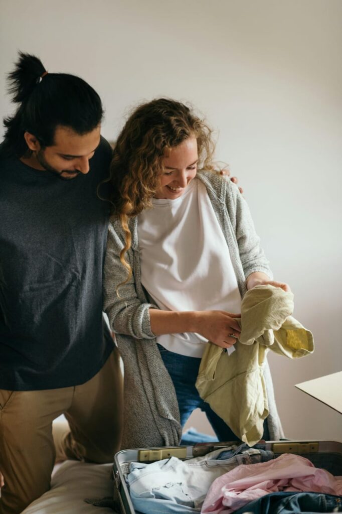 "Two people preparing for a trip, packing a suitcase on a bed, with one person holding a piece of clothing and various clothes inside the suitcase, symbolizing the excitement and anticipation of travel.