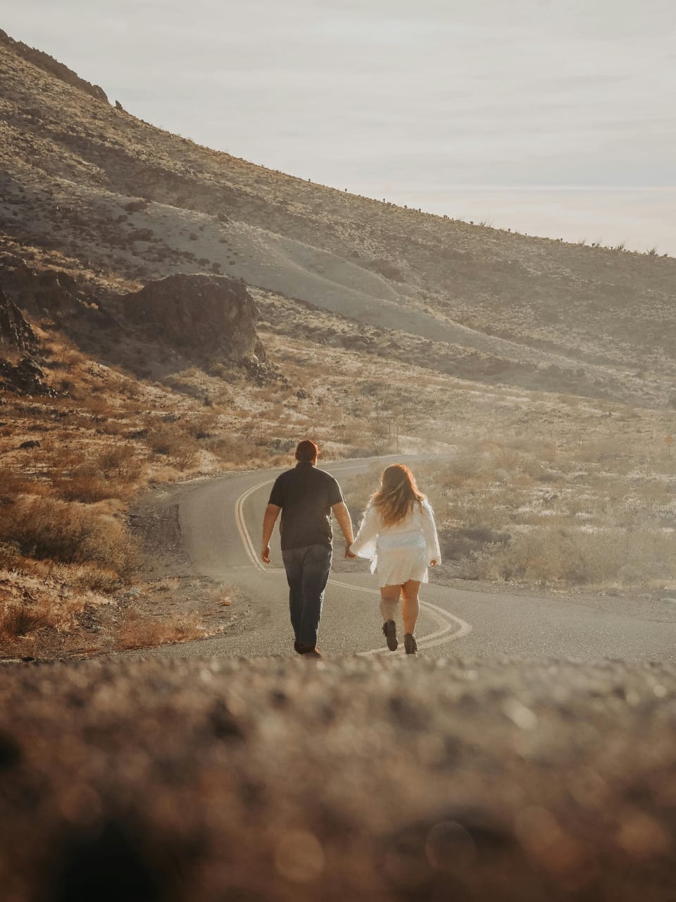 "A couple walking hand in hand on a winding road through a barren, rocky landscape with mountainous terrain and sparse vegetation, suggesting early morning or late afternoon lighting. The scene captures a sense of adventure and togetherness.